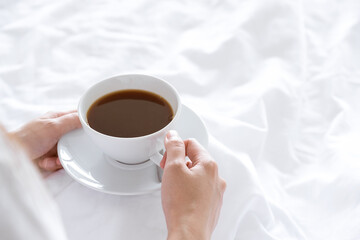 hands holding white cup of coffee on bedsheet in weekend with sun orange light. concept wake up in morning with warm drink to awake self in vacation or holiday relax time in bed room with copy space.