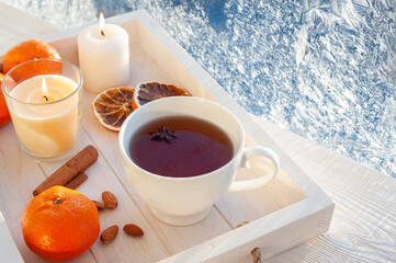 Winter tea at the frosty window. Tea with tangerines, cinnamon and nuts in a white cup on a white wooden tray.