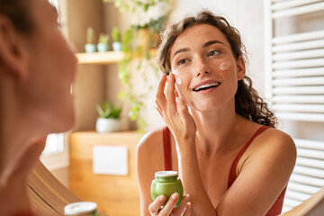 Woman applying moisturiser on face during morning routine - obrazy, fototapety, plakaty