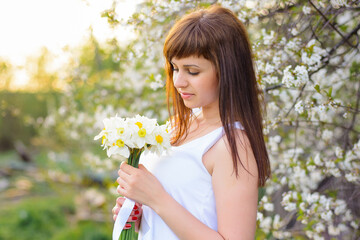 beautiful young girl with a bouquet of daffodils in a white dress on a background of cherry blossoms