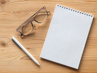 An open notebook with a blank sheet of paper on the wooden table next to the glasses and a white pen.