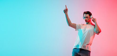 Music, drive. Young caucasian man's portrait on gradient blue-pink studio background in neon light. Concept of youth, human emotions, facial expression, sales, ad. Half length, copyspace. Flyer