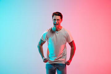 Crazy angry, screaming. Young caucasian man's portrait on gradient blue-pink studio background in neon light. Concept of youth, human emotions, facial expression, sales, ad. Half length, copyspace.