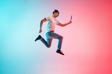 Fototapeta na wymiar Winner with tablet. Young caucasian man's jumping on gradient blue-pink studio background in neon light. Concept of youth, human emotions, facial expression, sales, ad. Full length, copyspace.