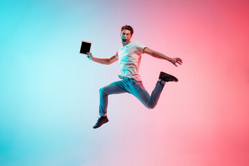 Obraz na płótnie Canvas Shouting with tablet. Young caucasian man's jumping on gradient blue-pink studio background in neon light. Concept of youth, human emotions, facial expression, sales, ad. Full length, copyspace.