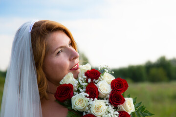 portrait of an attractive brown-haired woman in a veil with a beautiful smile and red lipstick, with a bouquet of red and white roses on a rural field, bride's bouquet