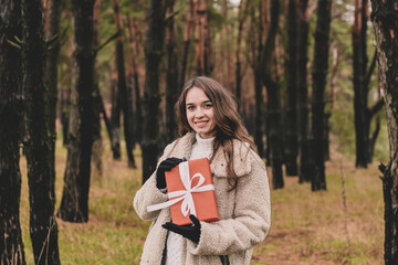 Beautiful young woman model appearance with long curly hair holding a Christmas present and smiling while being in a pine forest