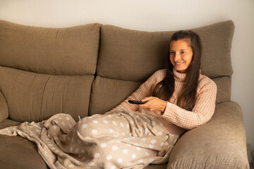 Woman lying on the sofa and covered with a blanket looks at the television with a smile and changes the channel with the remote control