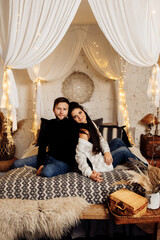 Obraz na płótnie Canvas Happy couple are sitting at the canopy bed, holding hands, smiling. Adorable young woman cuddle with charming attractive man at home. Loving husband celebrate christmas eve with beautiful wife