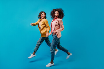 Fototapeta na wymiar Joyful female models running on blue background. Full-length portrait of two stylish women in jeans.
