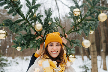 Happy smiling woman with fir tree antlers decorated with christmas ball held by her man. In a snowy winter forest.