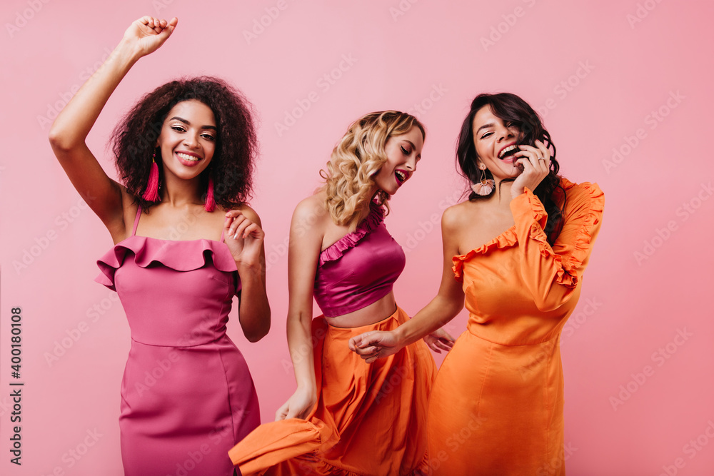 Wall mural African lady in pink dress dancing with friends. Blonde female model enjoying girl party.