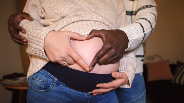 Interracial ethnic family relationship. An African-American man's hands with white girl's hand make a heart symbol of love on her expecting baby pregnant belly. Biracial couple.