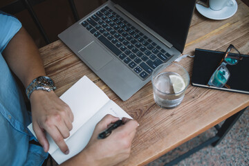 Crop faceless man writing in notepad at table with laptop