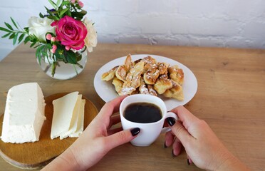 Cup of coffee in female hands, close-up. Homemade baked goods during quarantine.Homemade feta cheese on a wooden tabletop. Concept on the background of a white brick wall and a bouquet of roses.