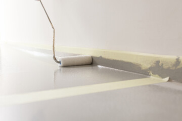 Close-up of a paint roller applying a new layer of grey paint on a floor, with the plinth protected with yellow painters tape.