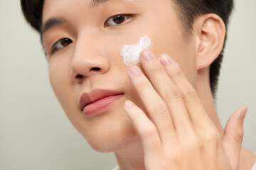 Skin care. Handsome young man applying cream at his face and looking at himself with smile while standing over white background