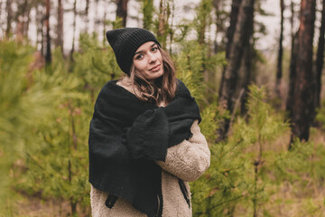 A beautiful young woman of model appearance with long curly hair in a sweater hat and a fur coat stands against the background of Christmas trees in a pine forest