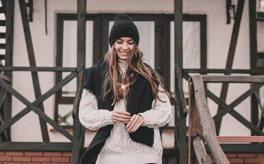 A beautiful young woman of model appearance with long curly hair in a sweater hat and a fur coat holds a sparkler in her hands and smiles against the background of a white private house