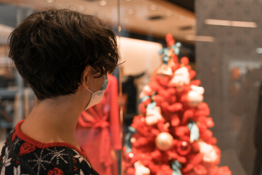 Shopping Woman Wearing Medical Mask At Mall Front Of Decorated Red Christmas Tree. Christmas And New Year Shopping. Gift Ideas. Ugly Christmas Sweater. 2021