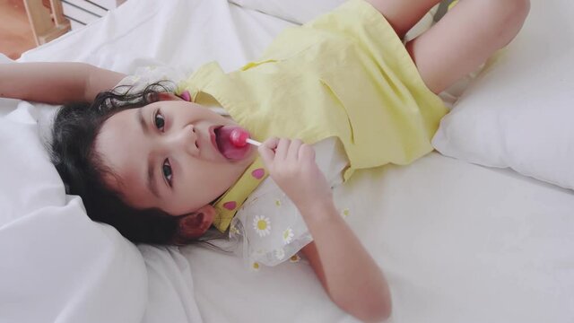 Asian little girl in a yellow dress was happily eating red lollipops and smiling on the white mattress and playing with the camera.