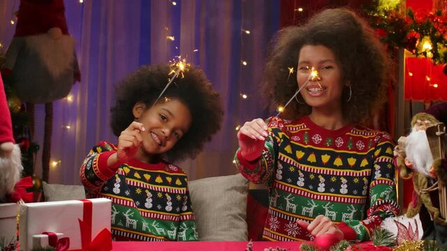 African American mom and little daughter in bright holiday sweaters light sparklers and paint a heart in the air. Happy family portrait, New Years celebration concept. Close up.