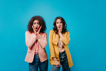 Portrait of two surprised women in colorful jackets. Indoor photo of shocked girls standing on blue background.