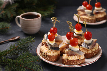 Canapes with salted herring, cheese, quail eggs and cherry tomatoes on rye croutons and cup of coffee on dark background. Horizontal format. Closeup