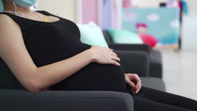 asian pregnant woman in medical face mask touching belly with care during waiting queue to meet doctor at kid hospital. 