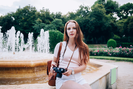 Portrait Of Serious Redhead Female 20s Traveler Resting On Fountain On Sightseeing Tour In City Using Camera For Taking Photos, Young Pretty Woman Enjoying Vacations Sightseeing On Journey