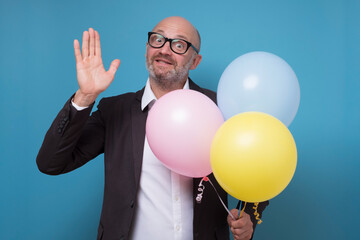 Mature caucasian man with balloons smiling happily and cheerfully, waving hand, welcoming and greeting you . Studio shot on blue wall.