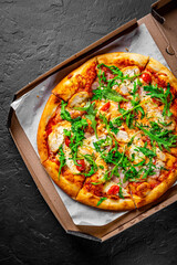 Pizza with chicken, bacon, cheese, tomato and arugula in paper box. Italian pizza on Dark grey black slate background