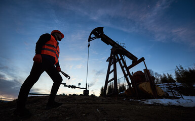 Low angle of male oil worker standing near petroleum pump jack under beautiful evening sky....