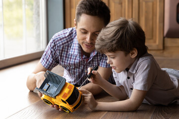 Little handyman. Focused son kid lying on warm wood floor fix toy car with help of young daddy....