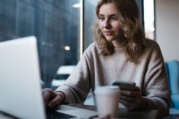 Focused young businesswoman with smartphone using netbook while working remotely in cafeteria