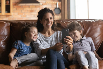 Millennial single mother spend time together with little son daughter at home enjoy family rest. Happy nanny and two small children use mobile phone take selfie together watch video at social media