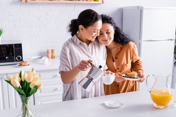 Hispanic same sex couple pouring coffee and holding pancakes near flowers on blurred foreground in kitchen