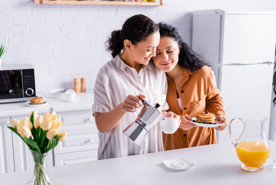 Hispanic same sex couple pouring coffee and holding pancakes near flowers on blurred foreground in kitchen
