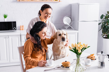 Happy hispanic lesbian couple looking at labrador near table with breakfast and tulips in kitchen...