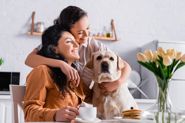 Cheerful lesbian hispanic woman hugging partner and labrador during breakfast in kitchen on blurred...