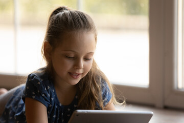 Cute little girl relax on warm floor by large window concentrated on screen of digital pad device play game online. Junior schoolgirl chat with friends classmates in social network watch funny cartoon