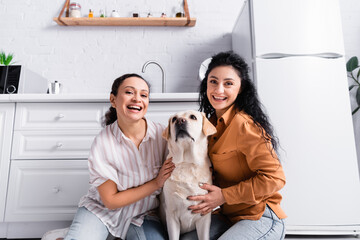 cheerful hispanic lesbian couple smiling at camera while embracing labrador dog in kitchen