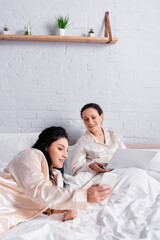 Fototapeta premium Hispanic lesbian woman showing smartphone to same sex lover using laptop in bed, blurred foreground
