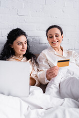 Smiling Hispanic woman holding credit card near partner with laptop on blurred foreground on bed