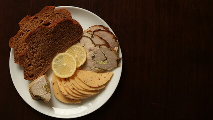 dark food background with a top view of a white platter with cold cuts of meat and cheese, slices of rye bread and lemon wedges with space for text on the right on a brown textured surface