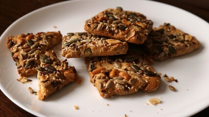 square cookies made from yeast-free dough sprinkled with seeds and nuts close-up on a white plate standing on a dark wooden table surface, healthy veggie natural home cooking snack
