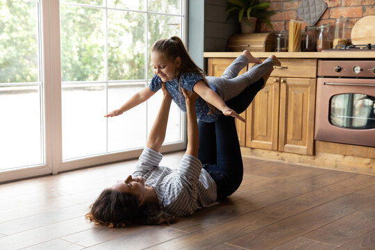 Cute Gymnastics. Happy Smiling Young Mom Supporting Little Daughter In Air Doing Flying Plane Exercise. Grown Elder Sister Playing Active Game With Younger Girl Lying On Heated Floor At Home Kitchen