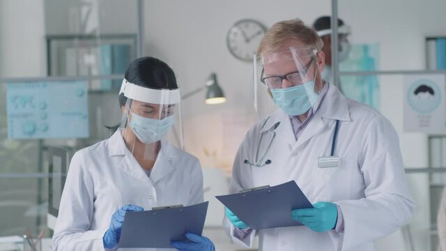 Young Asian female doctor and Caucasian male physician in lab coats, protective mask, screen and gloves writing on clipboard and having discussion while working in clinic during covid-19 pandemic