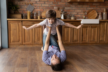 Active leisure with kid. Loving young daddy enjoy entertaining little son on weekend. Strong dad lie on wooden floor at kitchen interior hold small boy on outstretched arms play airplane flying in air