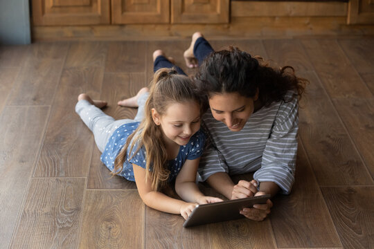 Funny learning. Happy small daughter and positive young mommy lie on warm wooden floor look at tablet screen engaged in shopping online. Little girl study language in education app with friendly nanny